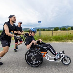 Young man being pushed during a race in the Joelette Finisher 3D Slider - Adjustable single-ski attachment for all-terrain wheelchairs, allowing for snow sports participation.