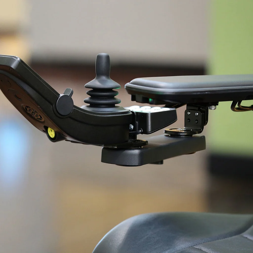Front view of the Retractable Joystick Mount on wheelchair. A sleek, metal joystick mount attached to a wheelchair armrest. The joystick can be easily swung outwards or retracted sideways for improved accessibility.